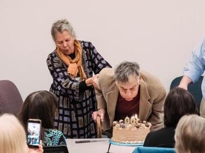 Mel bending over to blow out the candles on a 100th birthday cake as friends look on.