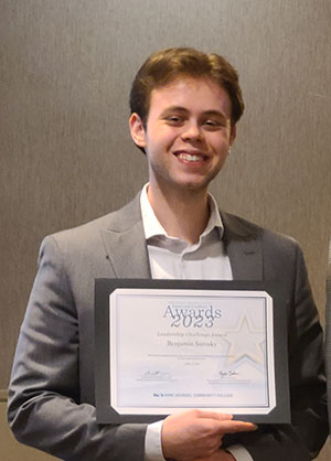 Ben holds up a certificate and smiles. He is dressed in a formal suit.