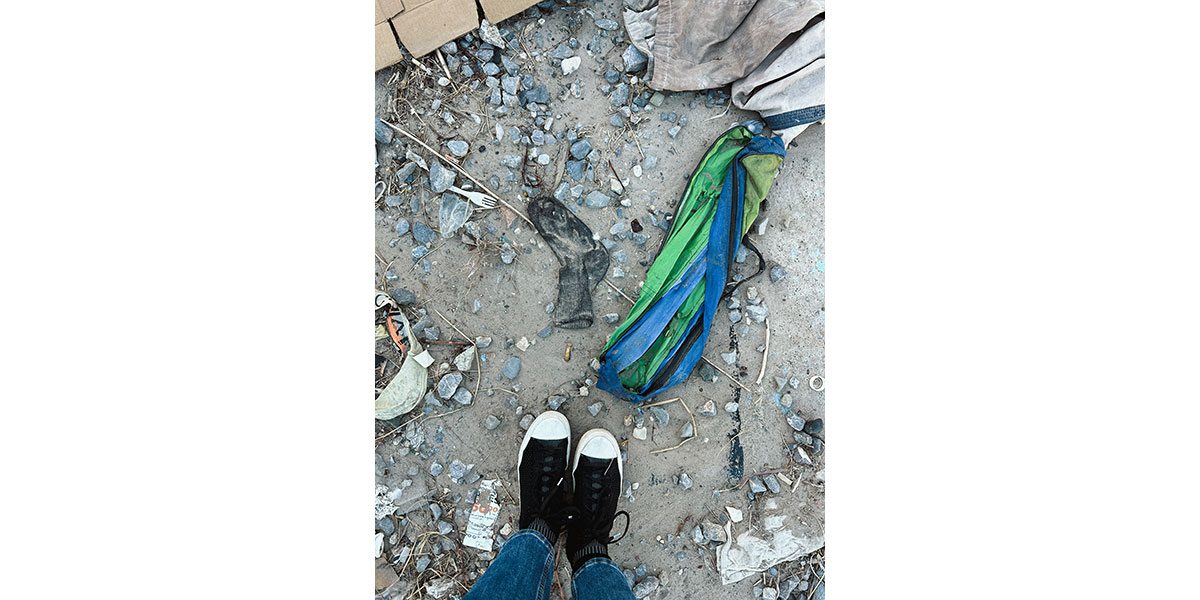 A person wearing black sneakers and blue jeans stands on gravelly ground littered with a dirty sock, green and blue material and various pieces of trash.