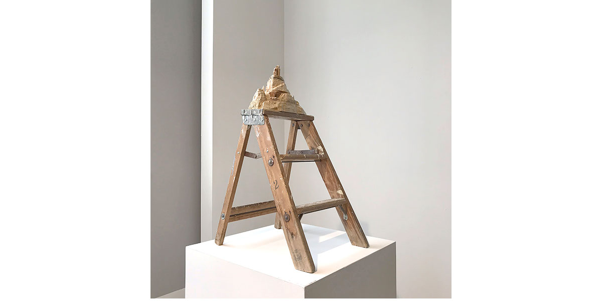 A wooden stepladder stands on a white pedestal in a gallery setting. Perched on the top step of the ladder is a small, tan-colored sculpture resembling a craggy mountain peak or a miniature rock formation.