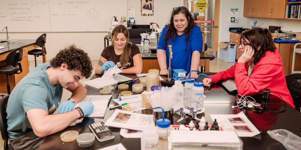 A teacher and group of students gather around a table full of science equipment.
