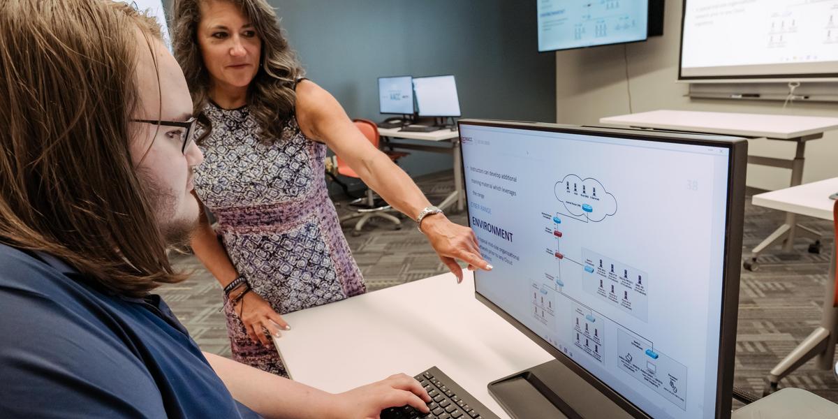 A female professor points to a computer screen while a student types on a keyboard and looks on.