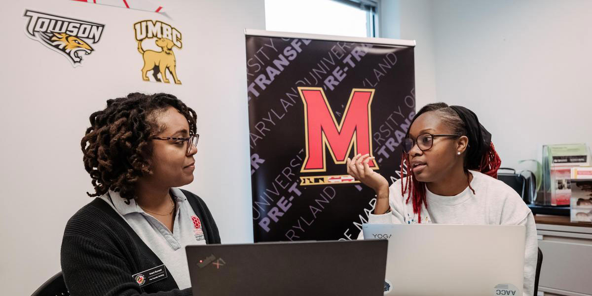 A transfer advisor speaking to an AACC student while graphics from local colleges hang on the wall.