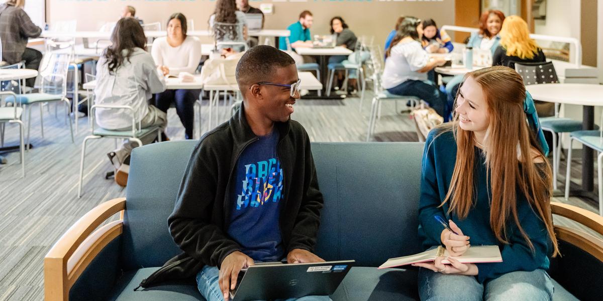 Students talking to each other on couch with students in background.