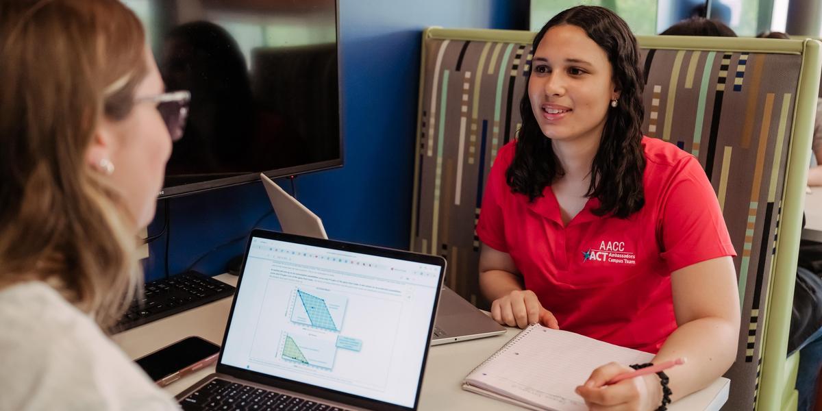 Two students sitting in a booth. One has a laptop and the other is writing in a notebook.